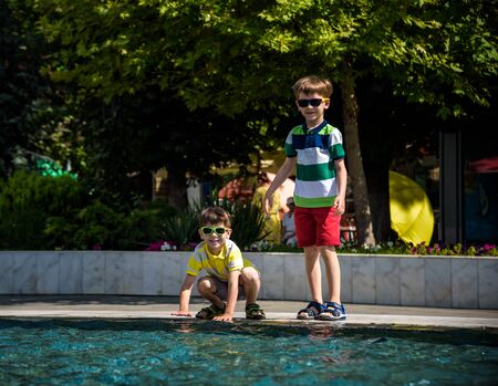 Group of happy children playing outdoors near pool or fountain. Kids having fun in park during summer vacation. Dressed in colorful t-shirts and shorts with sunglasses. Summer holiday concept.の写真素材