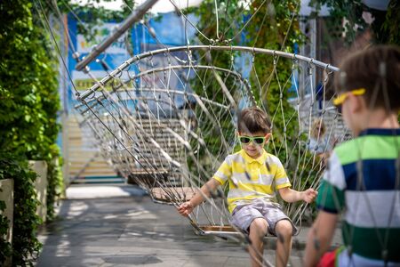 Little adorable kid sitting on chain swing during summer holidays on vacation. Boy relaxed and happy wearing casual clothes in sunglasses. Active summer leisure for kids in the city concept.の写真素材