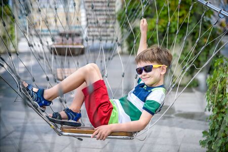 Little adorable kid sitting on chain swing during summer holidays on vacation. Boy relaxed and happy wearing casual clothes in sunglasses. Active summer leisure for kids in the city concept.の写真素材