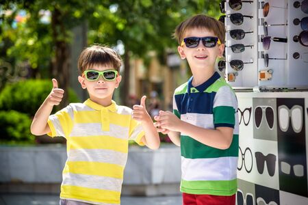 A child and a choice of sunglasses. Two little boys are standing in sun-proof glasses against the background of a shop window with glasses. Kid help choose each other. Sunglasses sale during summer vocation.の写真素材