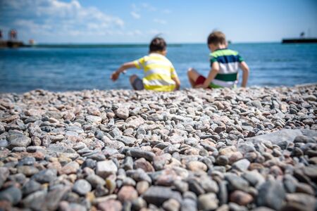 Two Caucasian baby boys is sitting at the beach. Kids in pants with his back sitting on the sea on a rocks pebbles. Sea vacations on with kids.の写真素材