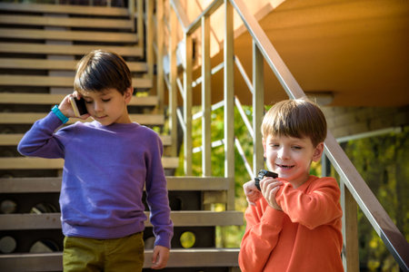 Two friends calling each other while standing on stairs outdoor. One boy has mobile phone, other has smart watch with GPS. Protection and safe communication with children concept.の写真素材