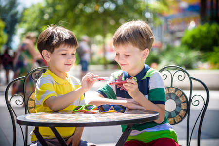 Two little kid boys waiting on table for healthy breakfast in hotel restaurant or city cafe. Child sit on comfortable chair play with toy aircraft, relaxed, enjoy their vacation. Summer holiday with children concept.の写真素材