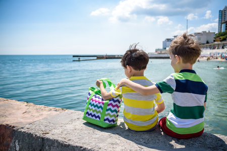 Two beautiful little boy sibling brothers in vest and shorts sitting on breakwater against the sea and shore in summer. Preschool children best friends enjoying summer vacations on sea. View from back.の写真素材