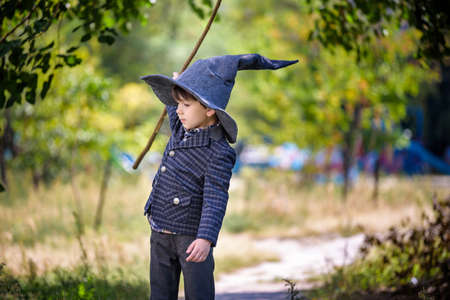 Toddler boy in pointed hat playing with magic wand outdoors. Little wizard. Halloween concept.の写真素材