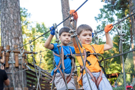 Two cute happy young children, boy and his brother in protective harness, carbine and safety helmets on rope way on bright sunny day on green foliage bokeh background. Outdoors activities, games concept.の写真素材