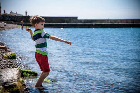 The little boy by the sea throws stones in water. Relaxation and vacation with kids. Happy childhood.の写真素材