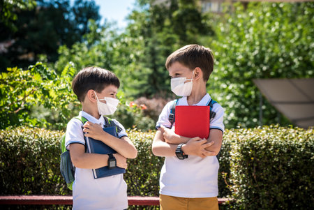 two boys, schoolchildren in black school uniforms with backpacks in a medical mask go to school independently, hold hands, friendship concept, study concept during coronavirus, back to school.の写真素材