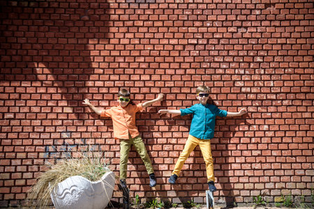 Two teenage boys sibling brothers with sunglasses in casual clothes are looking at camera and smiling, standing against yellow brick wall. Friendship concept.の写真素材