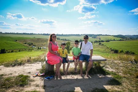 Happy family with two children holding hands of each other and sitting on bench with countryside background. Young couple of parents with kids enjoying nature together.の写真素材