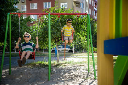 Two little kid boys having fun with swing on outdoor playground. Children, best friends and siblings swinging on warm sunny spring or autumn day. Active leisure with kids. Casual boy fashion.の写真素材