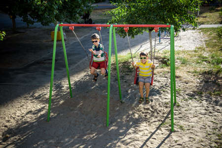 Two little kid boys having fun with swing on outdoor playground. Children, best friends and siblings swinging on warm sunny spring or autumn day. Active leisure with kids. Casual boy fashion.の写真素材