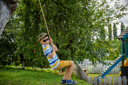 Happy little boy is having fun on a rope swing which he has found while having rest outside city. Active leisure time with children.の写真素材