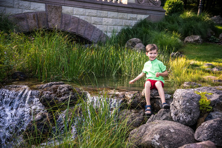 A young boy sits on the river bank, dreams. Warm summer or spring day. Cute Caucasian kid play with water.の写真素材