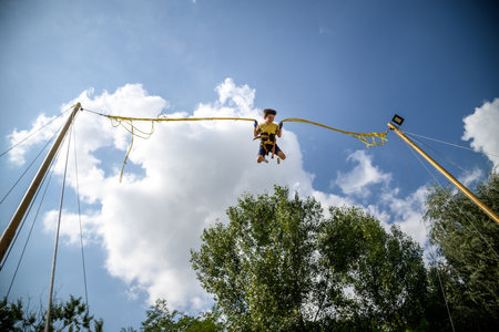 The boy is jumping on a bungee trampoline. A child with insurance and stretchable rubber bands hangs against the sky. The concept of happy childhood and games in the amusement park.の写真素材