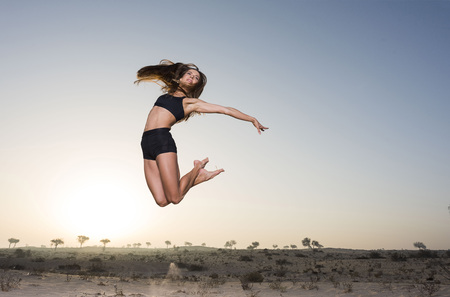 Beautiful athletic white girl in desert wearing black shorts and sports bra does a happy jump at sunrise or sunsetの写真素材