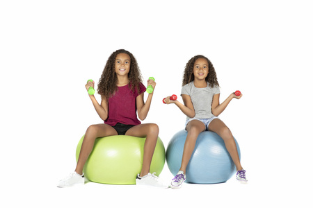 Two young playful children or pre teen biracial girls with brown curly hair playing or exercising on large rubber balls lifting dumbbellsの写真素材