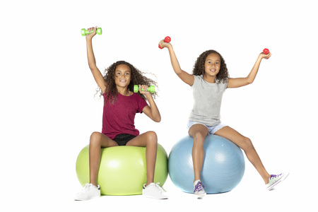 Two young playful children or pre teen biracial girls with brown curly hair playing or exercising on large rubber balls lifting dumbbellsの写真素材