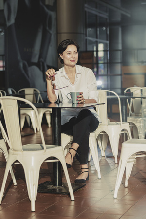 Beautiful European caucasian woman sitting in an outdoor cafe with sunglasses enjoying drinking a cup of coffee wearing a white shirt and black tight pants with a light flare in the backgroundの写真素材
