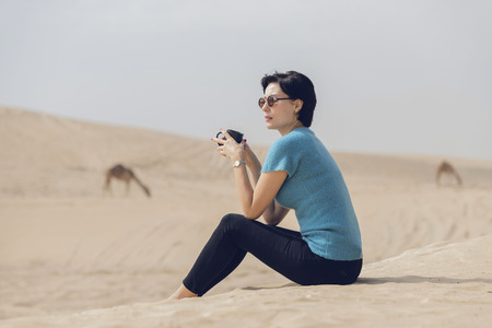 Beautiful European caucasian woman sitting on the sand dune in the desert with sunglasses relaxing drinking a cup of coffee with camels  grazing in the background.の写真素材