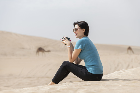 Beautiful European caucasian woman sitting on the sand dune in the desert with sunglasses relaxing drinking a cup of coffee with camels  grazing in the background.の写真素材