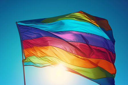 Rainbow flag waving in the wind against blue sky with sun raysの素材