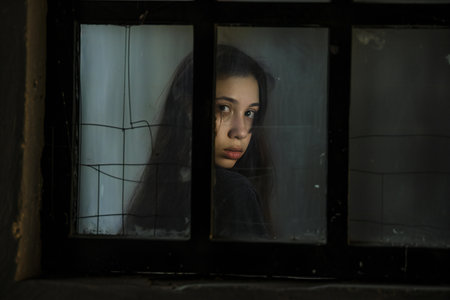 Young woman looking through the window in a dark room with dirty glassの素材