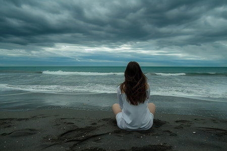 Young woman sitting on the beach and looking at the sea with cloudy skyの素材