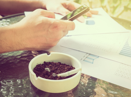A businessman is seriously working with mobile phone looking at graph paper with cigarette butt in ashtray, selective focus of business and financial working concept.の写真素材