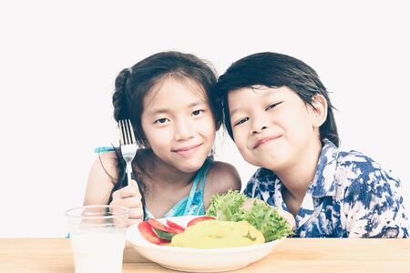 Vintage style photo of asian boy and girl are happily eating fresh vegetable salad with a glass of milk isolated over white backgroundの写真素材