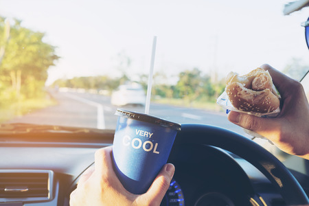 Man is dangerously eating hot dog and cold drink while driving a carの写真素材