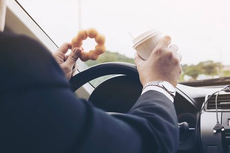 Man eating donuts with coffee while driving car - multitasking unsafe driving conceptの写真素材
