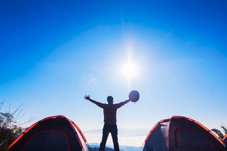 Man happy camping stand and raise his hand holding his hat on the mountain in the morning time during his trekking camp near to two small tents with sun cloud and blue sky background, Thailandの写真素材