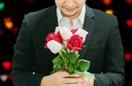 Man giving red and white bouquet roses to somebody in valentine day or other love festival event with black and blur colorful heart bokeh backgroundの写真素材