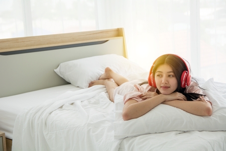 Portait of smiling lady in pink pajama wake up listening to music with headphone welcome morning light on the bedの写真素材
