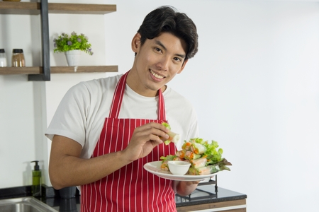 Young good looking man in red apron presenting the healthy menu of salad roll with smile and welcome people to taste, prepared in home kitchenの写真素材
