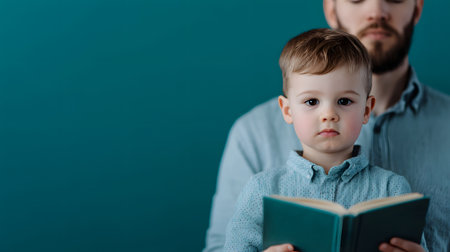 A young boy holding a book looks thoughtfully into the camera, while his father stands behind him. The background is a solid teal color, radiating warmth and connection in their moment.の素材