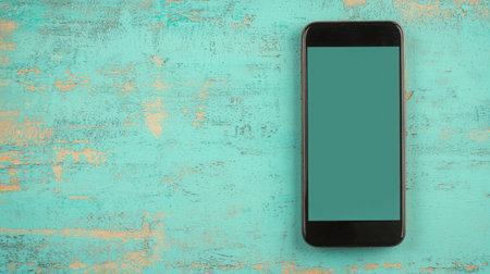 A sleek smartphone sits on a rustic turquoise wooden background, featuring a blank screen ideal for displaying various digital content or advertisements.の素材