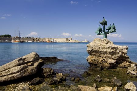Mandraki port in Rhodes Town, Greece, with dolphin statue in the foreground and St Nicholas fortress in the backgroundの写真素材