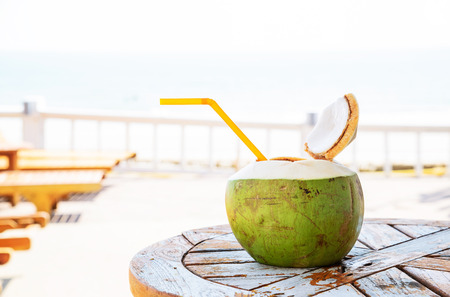 Coconut juice with straw on wood table against blurred beach background.