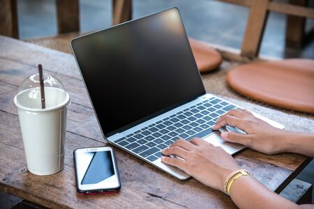 Young hipster woman's hands busy using her laptop on wooden table with smart phone in a coffee shop...
