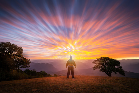 Silhouette of a man standing on top the hill and observes the idyllic sunrise. Male tourist hiking in the mountains stops to see the picturesque sunrise.の写真素材