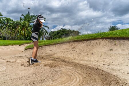Side view of golfer female shooting golf ball from sand bunker. Golfer hitting golf ball with sand blast.の写真素材