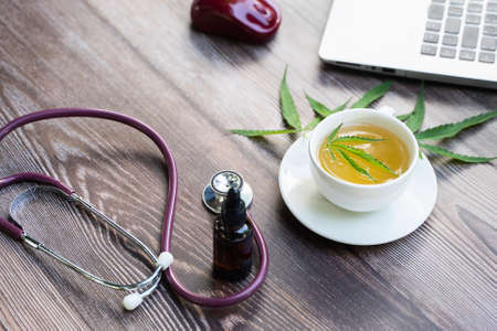 Cannabis oil in tea cup, Stethoscope, Laptop, leaf of cannabis and a bottle of CBD Hemp Oil on desk. Medical marijuana concept. Top view, flat lay of CBD oil hemp productsの写真素材