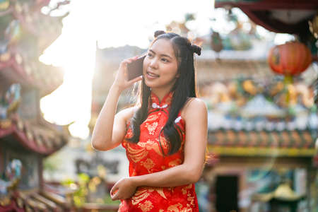 Portrait beautiful Asian young woman wearing red traditional Chinese cheongsam decoration and using smartphone for Chinese New Year Festival at Chinese shrineの写真素材