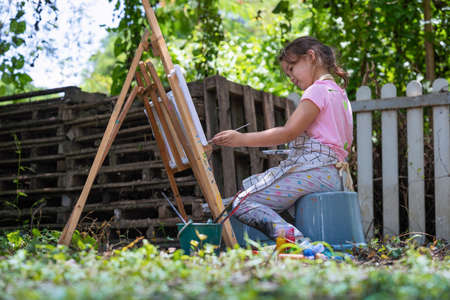 Little girl artist drawing a picture on canvas on an easel in nature, Cute little girl painting picture with a brush and color in backyard.  concept of art, hobby and relaxの写真素材