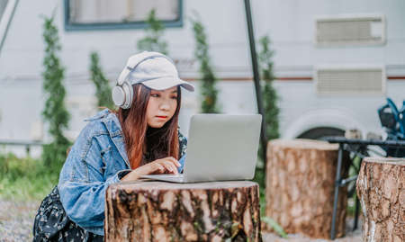 Young girl using laptop with caravan van behind, Relaxing in nature with Caravan trailer in the evergreen pine forest. Concept of leisure activity, eco tourism, camping siteの写真素材