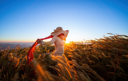 Woman in a hat standing and waving red cloth against the wind with sunset. A young girl stands against the background of the sunset in the grass field and admires the light sunの写真素材