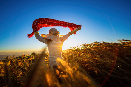 Woman in a hat standing and waving red cloth against the wind with sunset. A young girl stands against the background of the sunset in the grass field and admires the light sunの写真素材