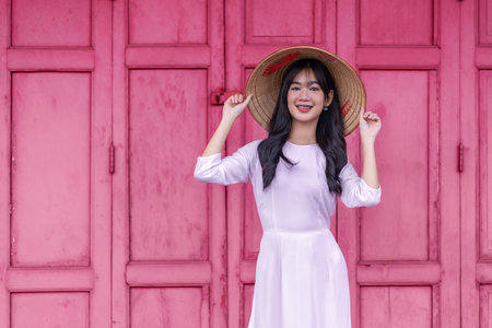 A beautiful Vietnamese schoolgirl in a white pink ao dai and pink door background.The Ao dai ( long-dress Vietnamese) is traditional costume of Vietnamese woman.Vietnamese young lady in traditional robeの写真素材